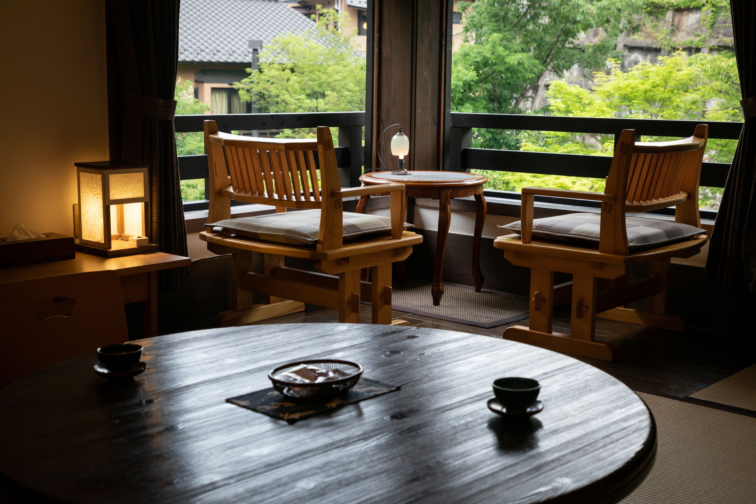 A traditional Japanese room with tatami flooring, a round wooden table, two wooden chairs, a small table with a lantern, and a view of greenery outside.