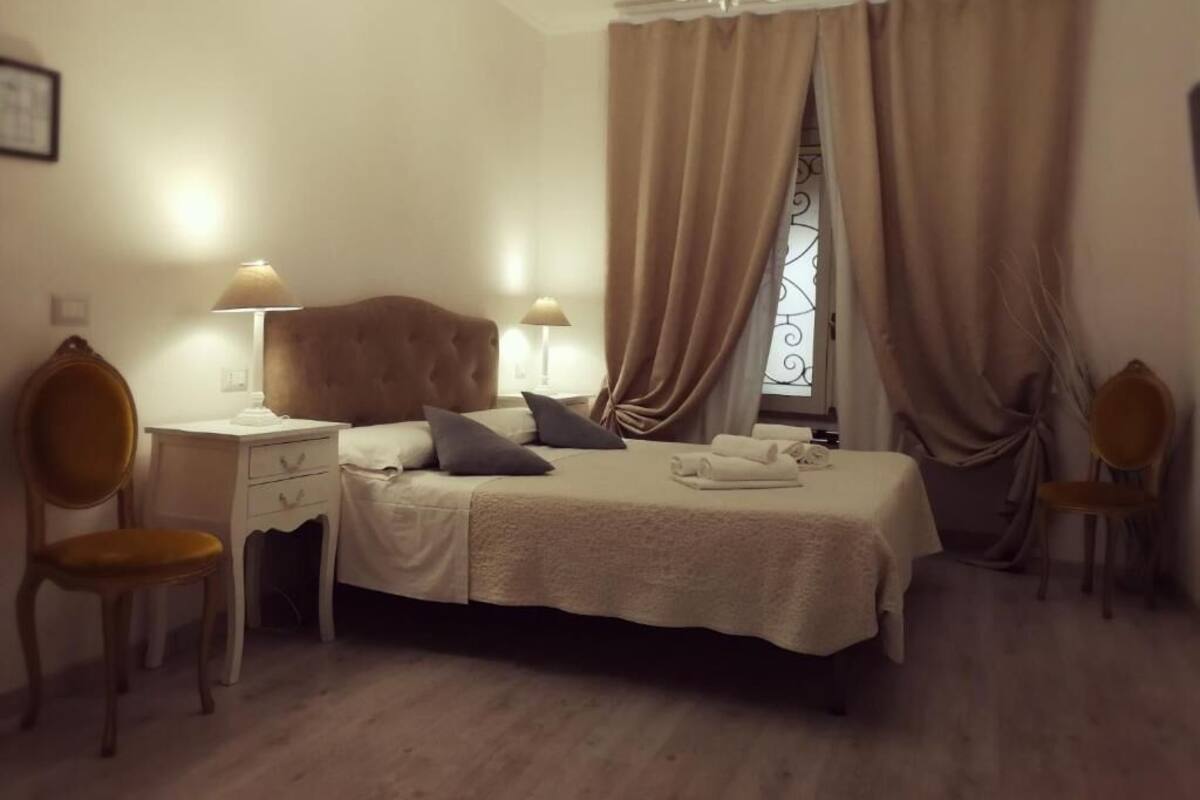 A bedroom with a bed, two bedside tables, a desk, two chairs, and a window with curtains.