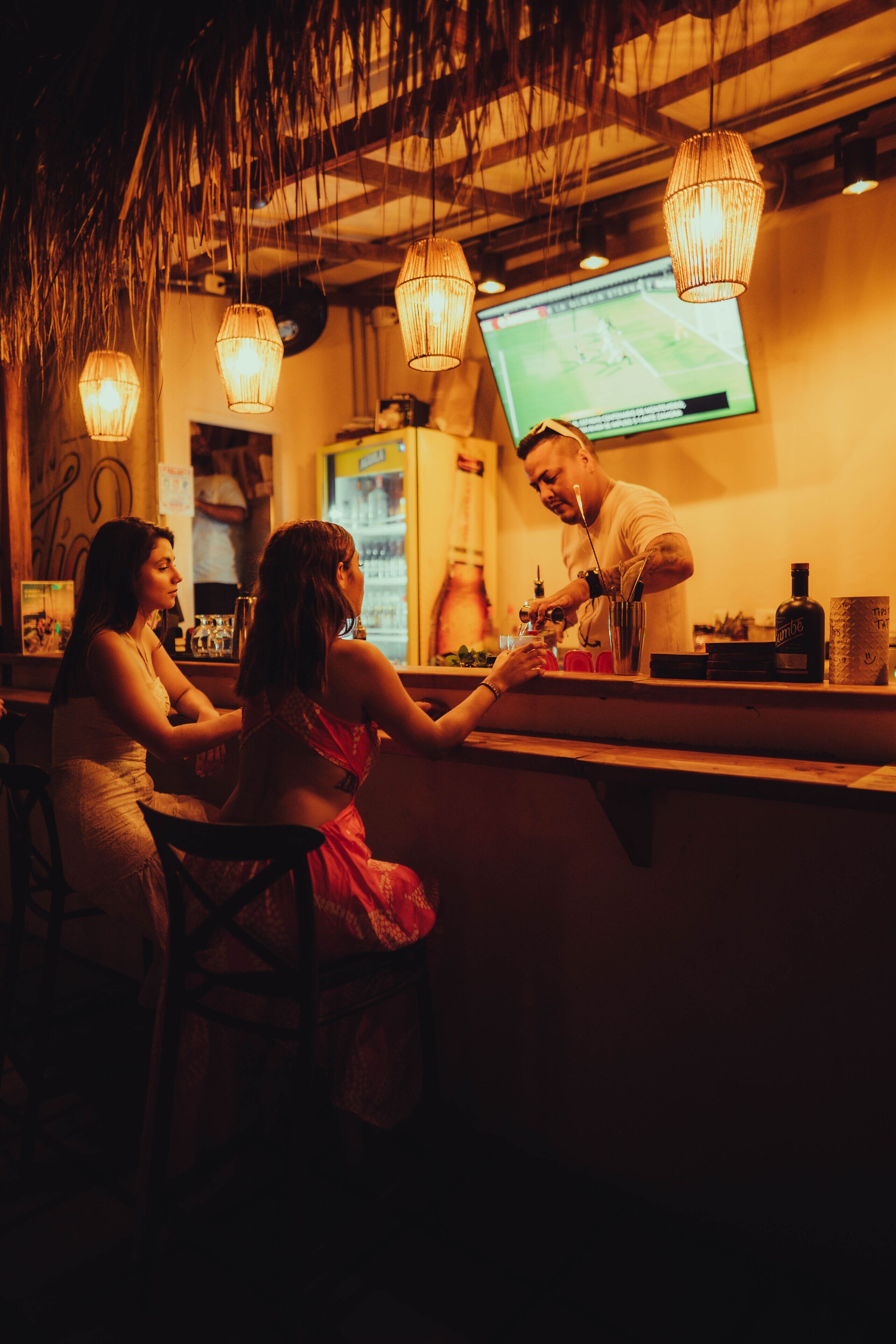 A bar area with hanging lights, a TV screen, and a bartender preparing drinks.