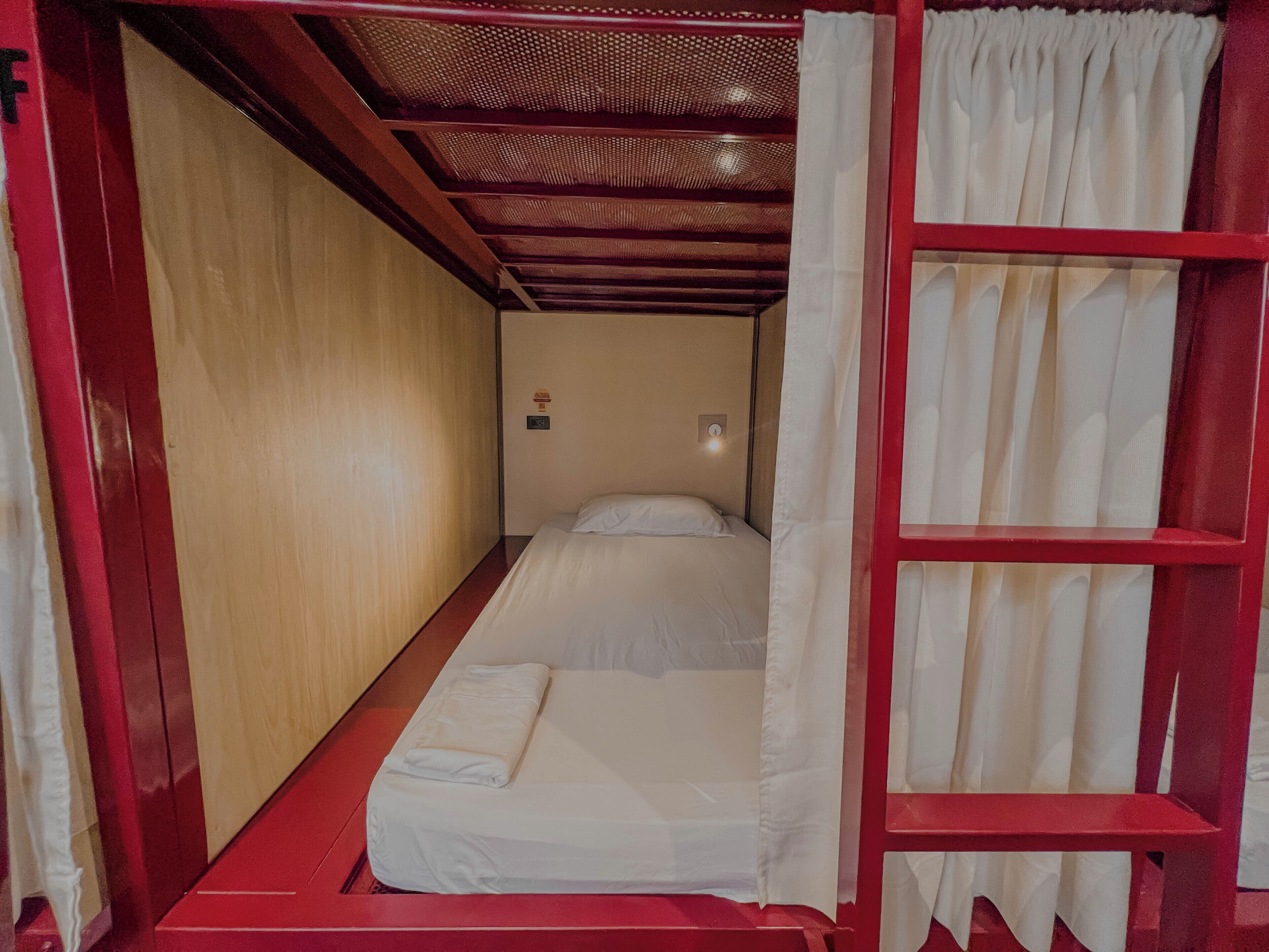 Een eenpersoonskamer met een houten vloer, een rode bedomgeving en een witte spreidekker.