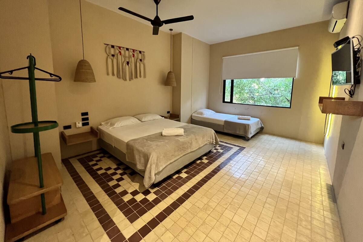 Ein Schlafzimmer mit einem Bett, einem kleineren Bett, einem Standventilator, einem Fenster und einem an der Wand befestigten Regal.