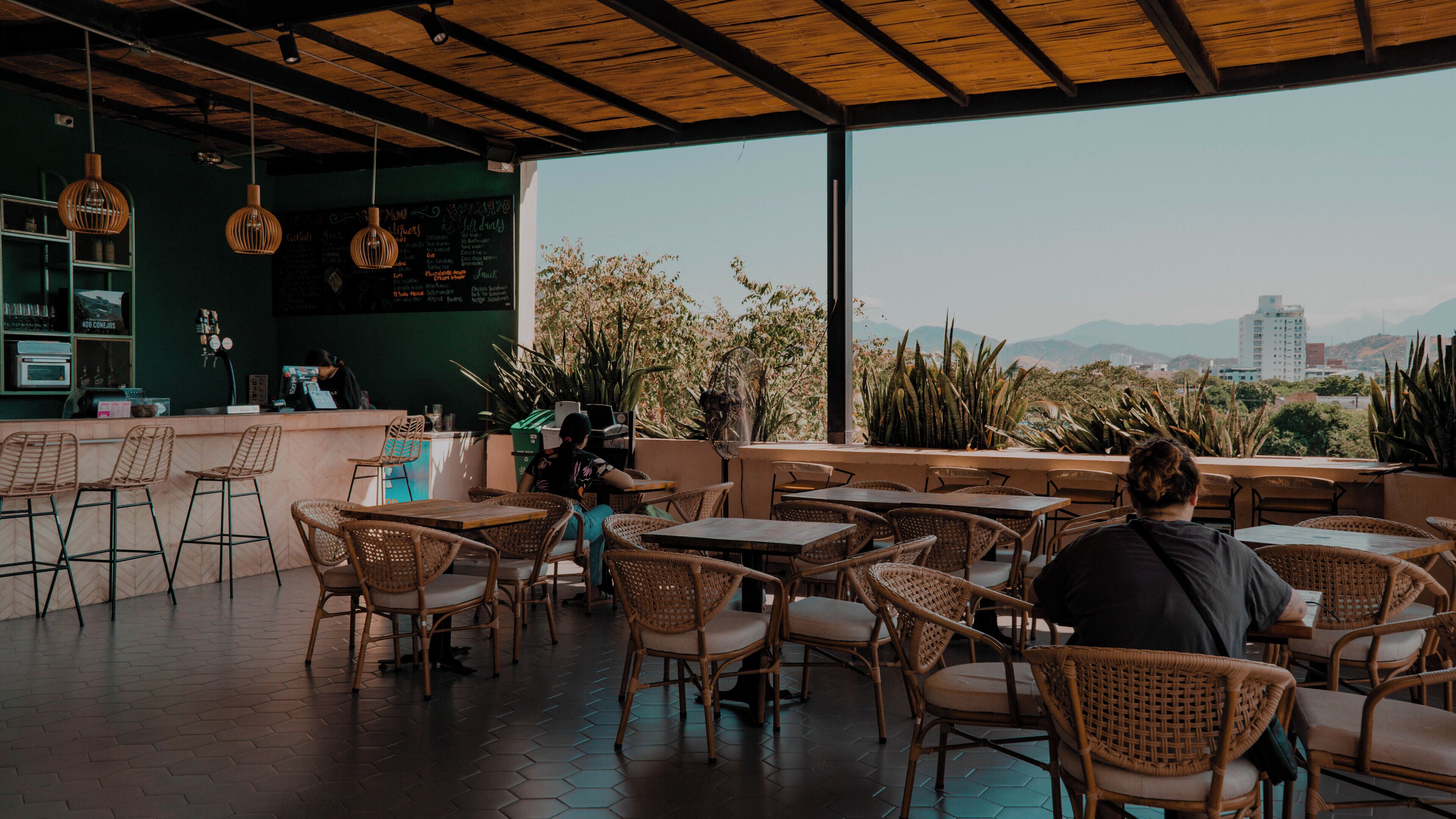 Un café al aire libre con mobiliario de mimbre, una zona de bar y vistas panorámicas de la ciudad.