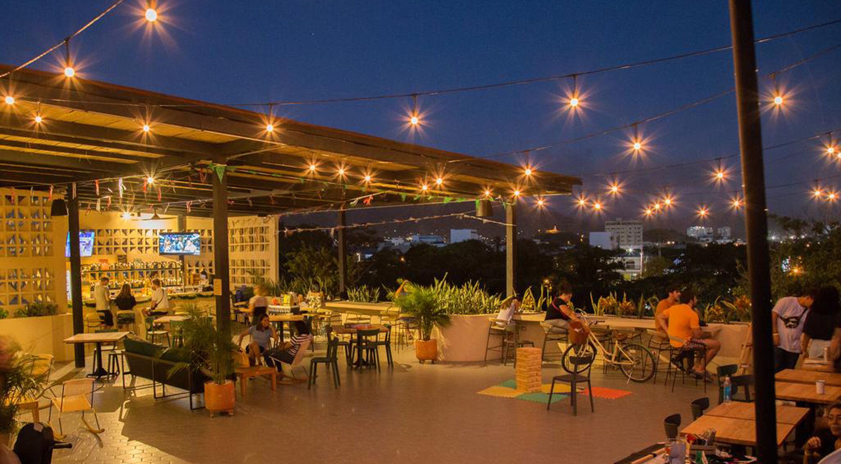 Uma área de estar ao ar livre com mesas e cadeiras, luzes de festa e vista para o horizonte da cidade.