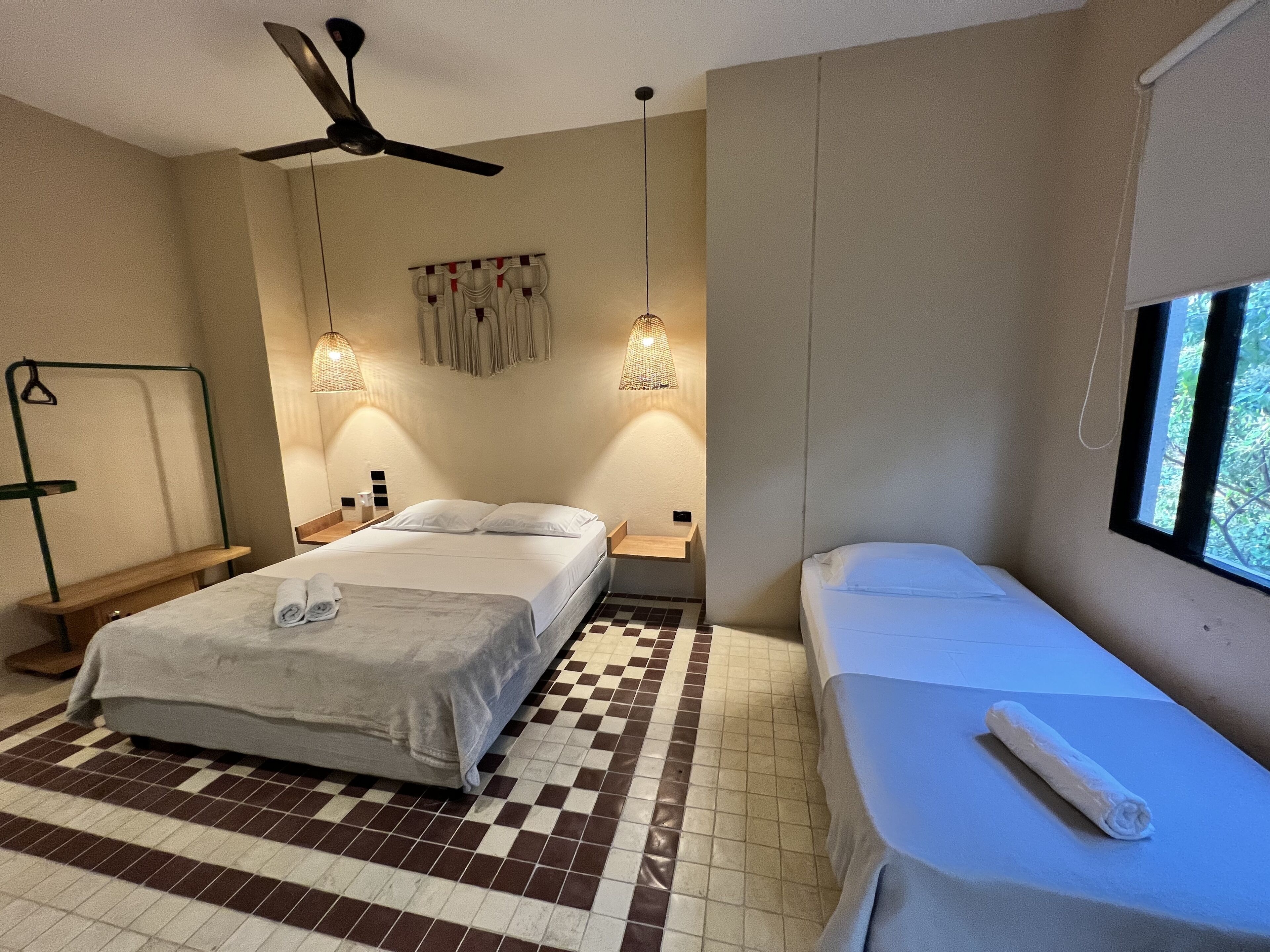A hotel room with a bed, a smaller bed or daybed, a standing lamp, a ceiling fan, and a window with a view of greenery.