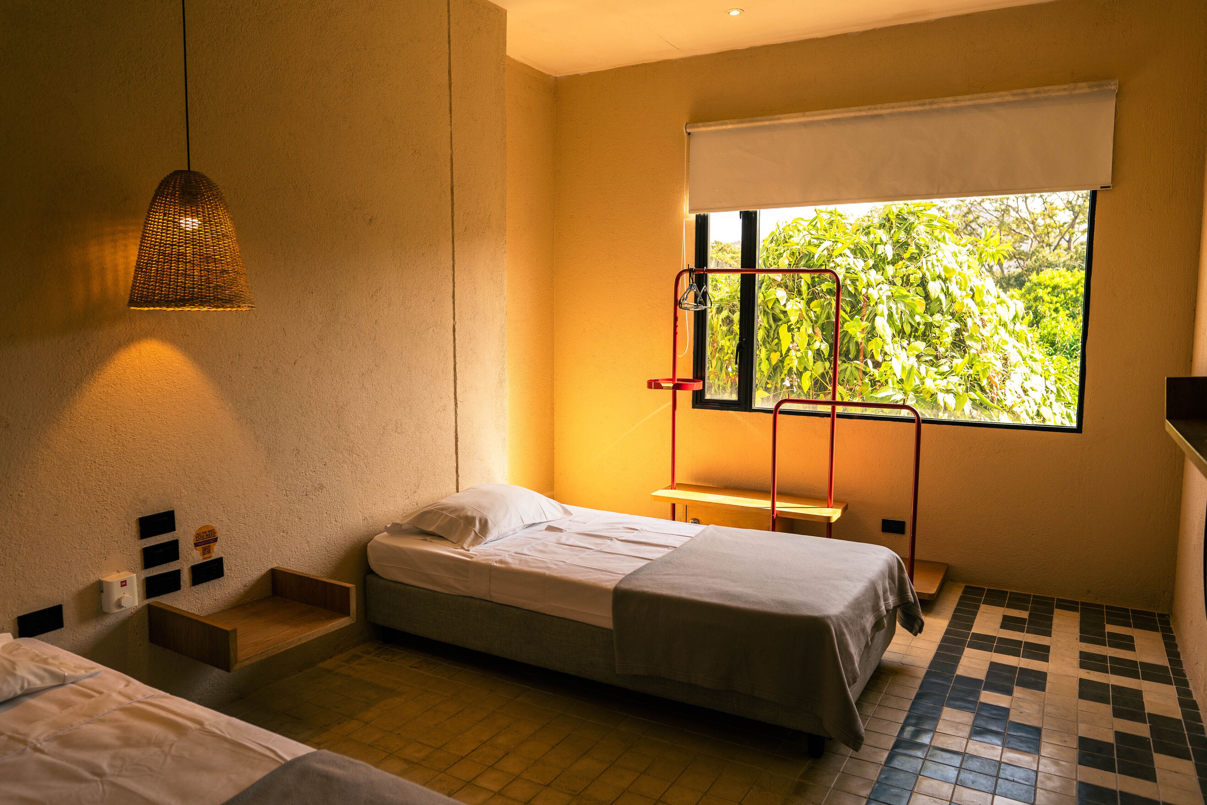 A small, modern room with a single bed, a bedside table, a hanging light fixture, a window with a view of greenery, and a tiled floor.