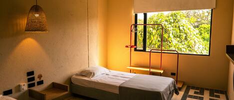 A small, modern room with a single bed, a bedside table, a hanging light fixture, a window with a view of greenery, and a tiled floor.