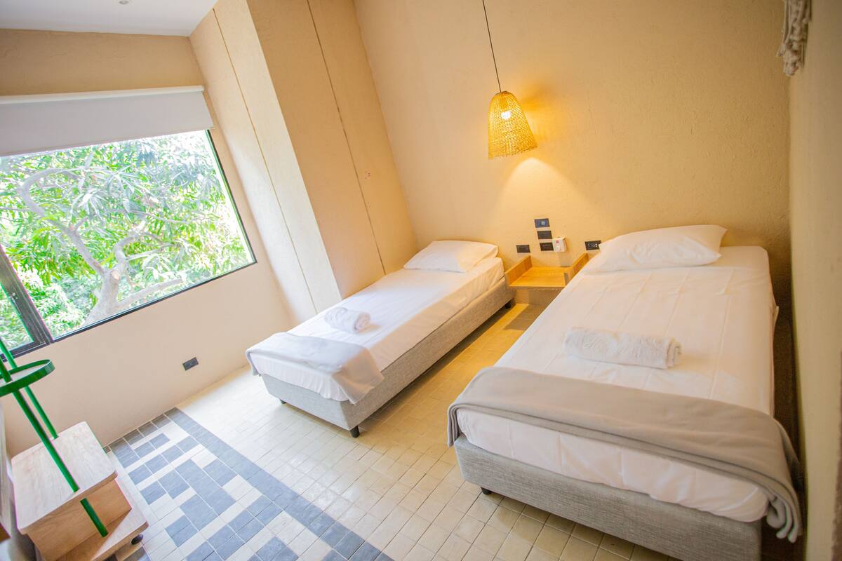 Ein Hotelzimmer mit zwei Einzelbetten, einem kleinen Tisch, einem Stuhl und einem großen Fenster mit Blick auf Grünflächen.