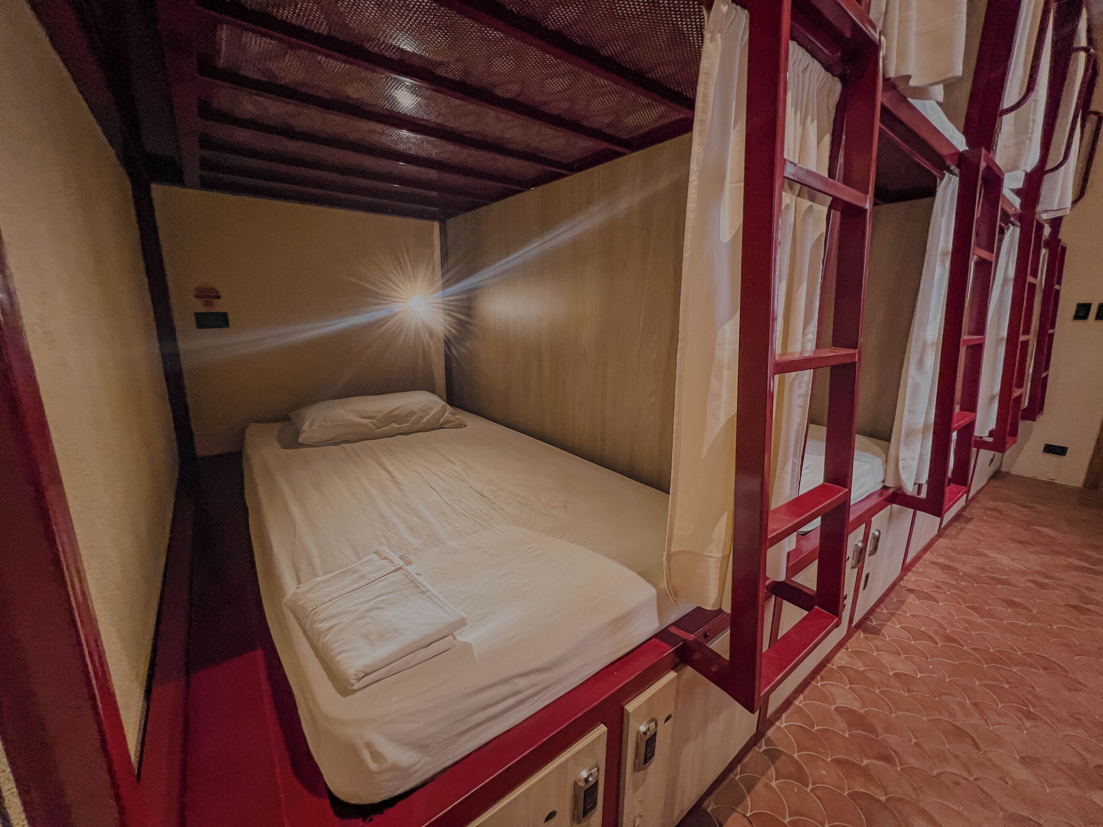 Una cama individual con estructura de madera y colchón, un pequeño mueble debajo y una ventana con cortinas.