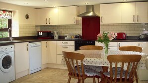 A compact kitchen with a washing machine, microwave, stove, and a dining area with a table and chairs. - Callestock Courtyard (Truro)