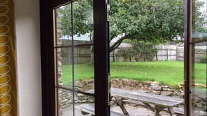 A view through a glass door into a backyard with a stone wall, a wooden picnic table, and a grassy area with trees. - Callestock Courtyard (Truro)
