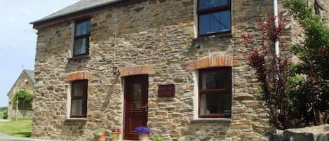 A stone building with a central entrance, two windows on the upper floor, and a small porch area with potted plants.