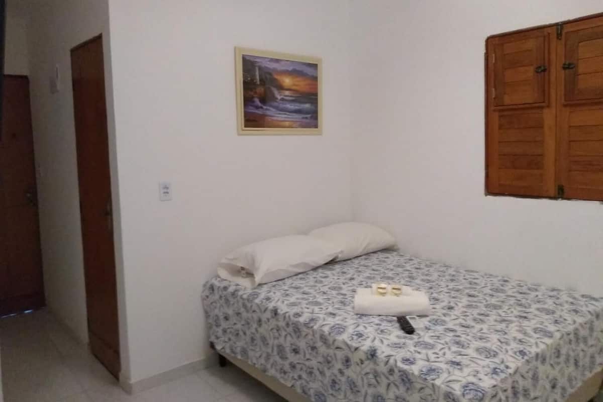 A small, single-bed room with a floral-patterned bedspread, a white pillow, and a small table with a towel and two bottles.