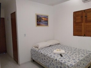 A small, single-bed room with a floral-patterned bedspread, a white pillow, and a small table with a towel and two bottles. - Pousada Praia Bela Maceio (Maceió)