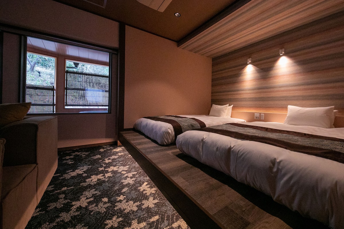 Une chambre d’hôtel moderne avec un grand lit, un canapé, une fenêtre donnant sur une clôture et une tête de lit en bois.