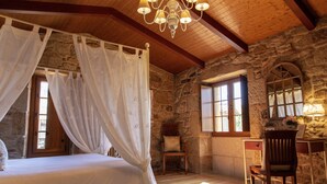 Un dormitorio con cama con dosel, pared de piedra, vigas de madera, un escritorio con silla y una ventana con vistas.
