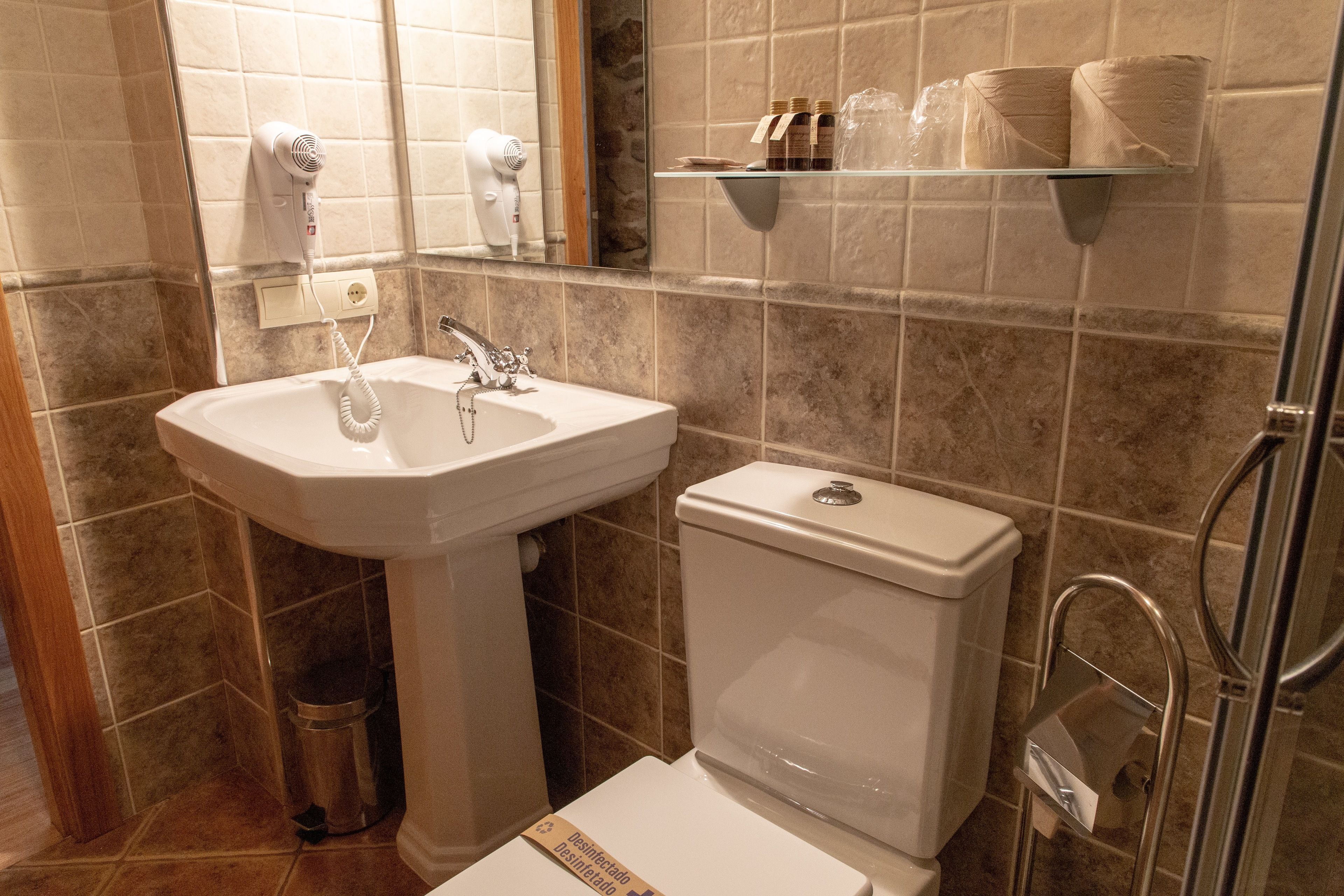 A bathroom with a pedestal sink, a toilet, and a glass shelf above the toilet holding toiletries.