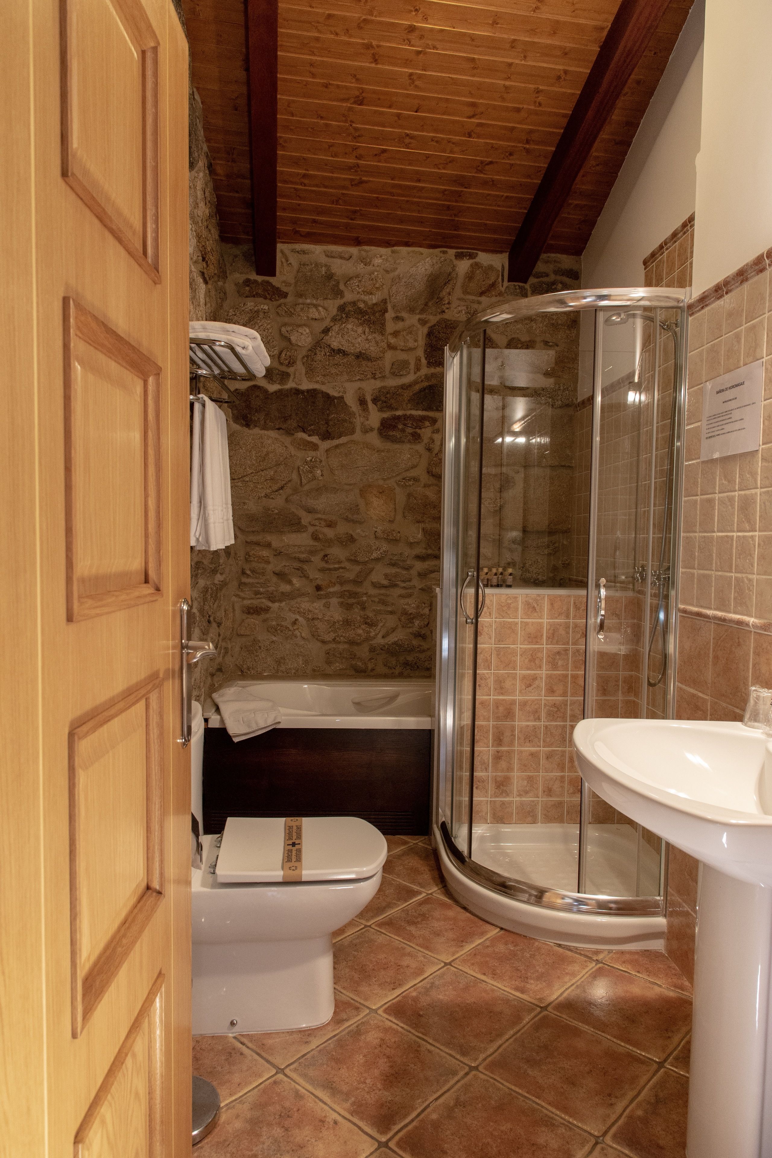 Une salle de bain avec un mur en pierre, une porte en bois, une douche en verre, des toilettes blanches et un lavabo.