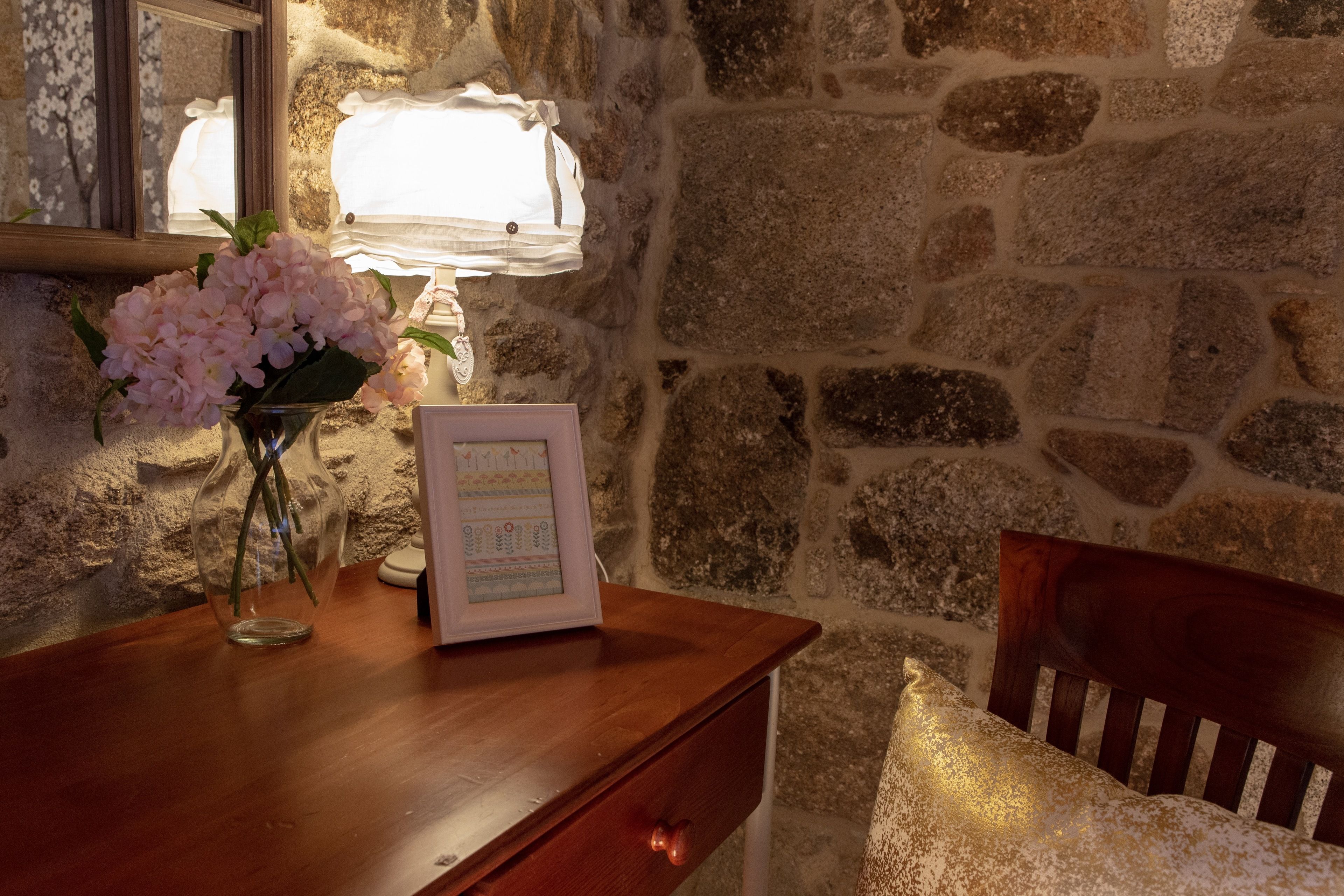 A wooden desk with a vase of flowers, a framed picture, and a lamp against a stone wall.