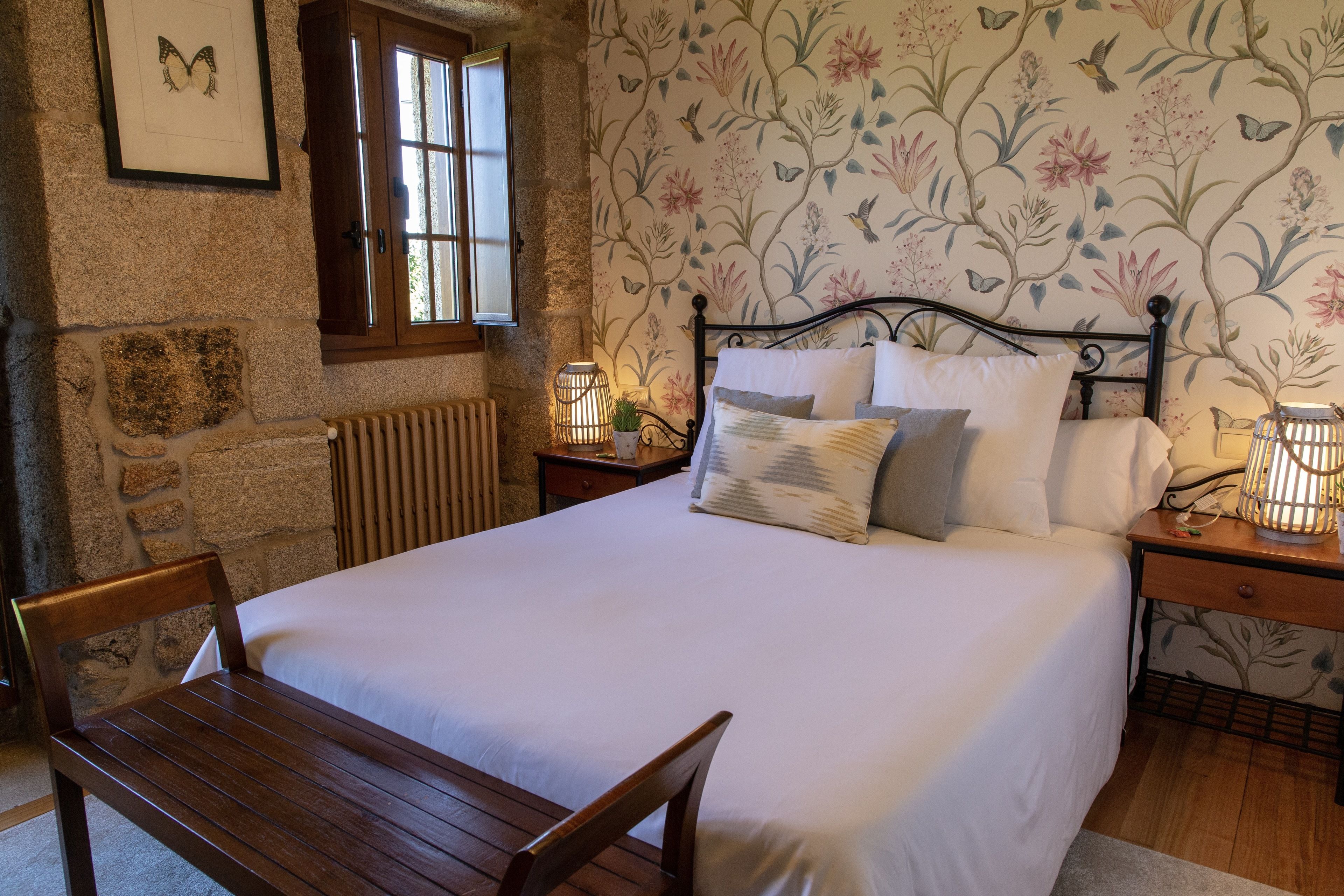 Quarto com cama grande, cabeceira de madeira, mesa de cabeceira com abajur, um banco de madeira e parede de pedra com janela.