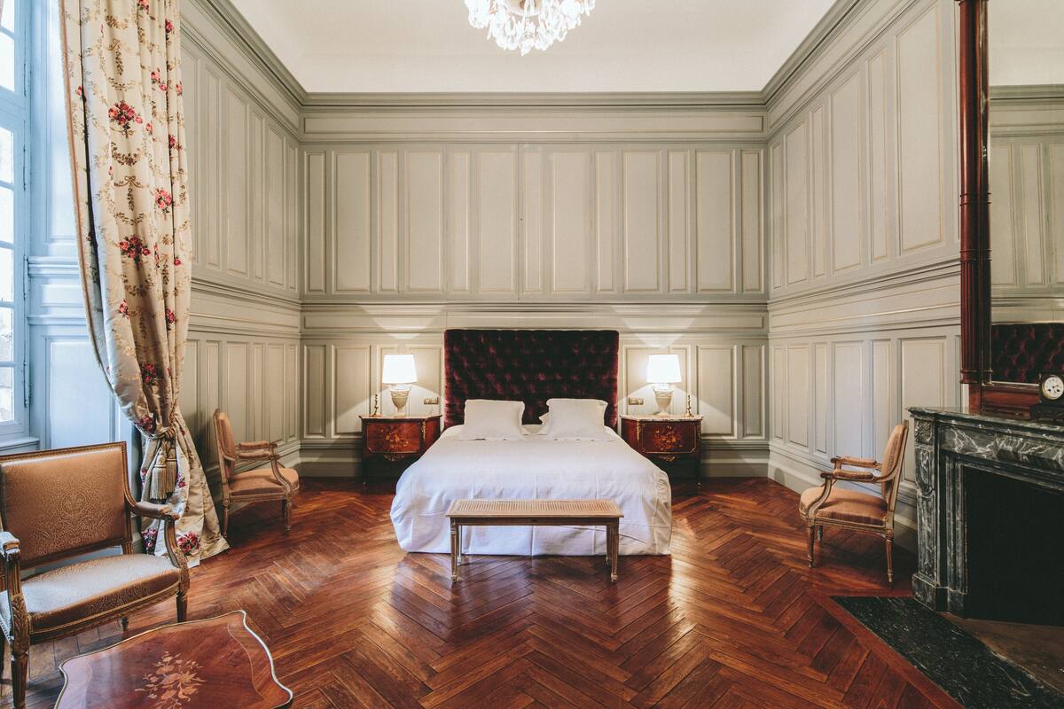 A spacious bedroom with a large bed, two bedside tables with lamps, a wooden bench, and a fireplace.