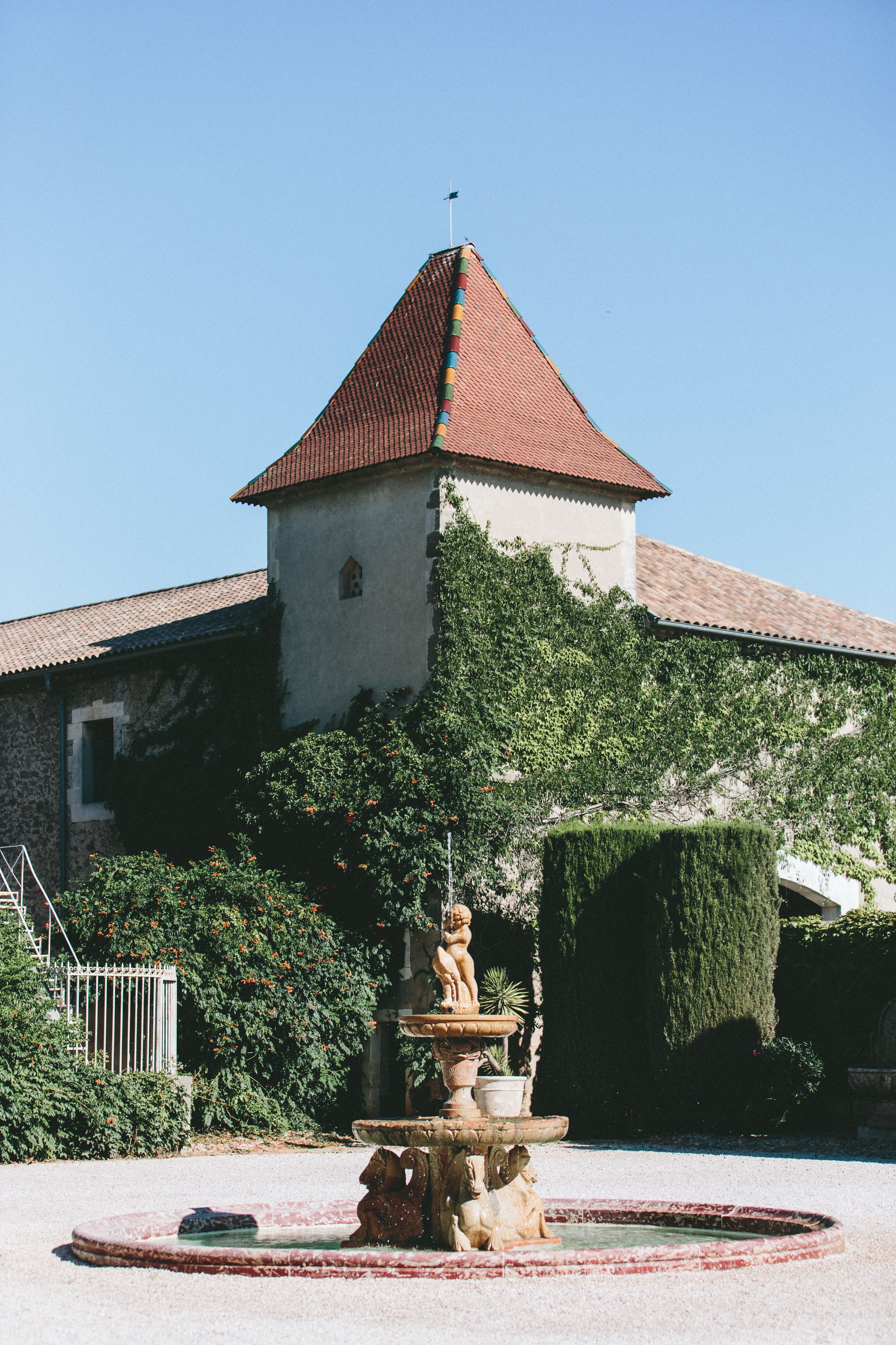 A historic building with a red-tiled roof, a stone facade, and a fountain with statues in the foreground.