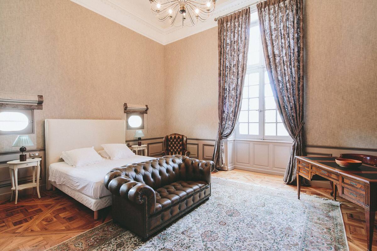 A bedroom with a large bed, a leather sofa, a wooden desk, and a chandelier.