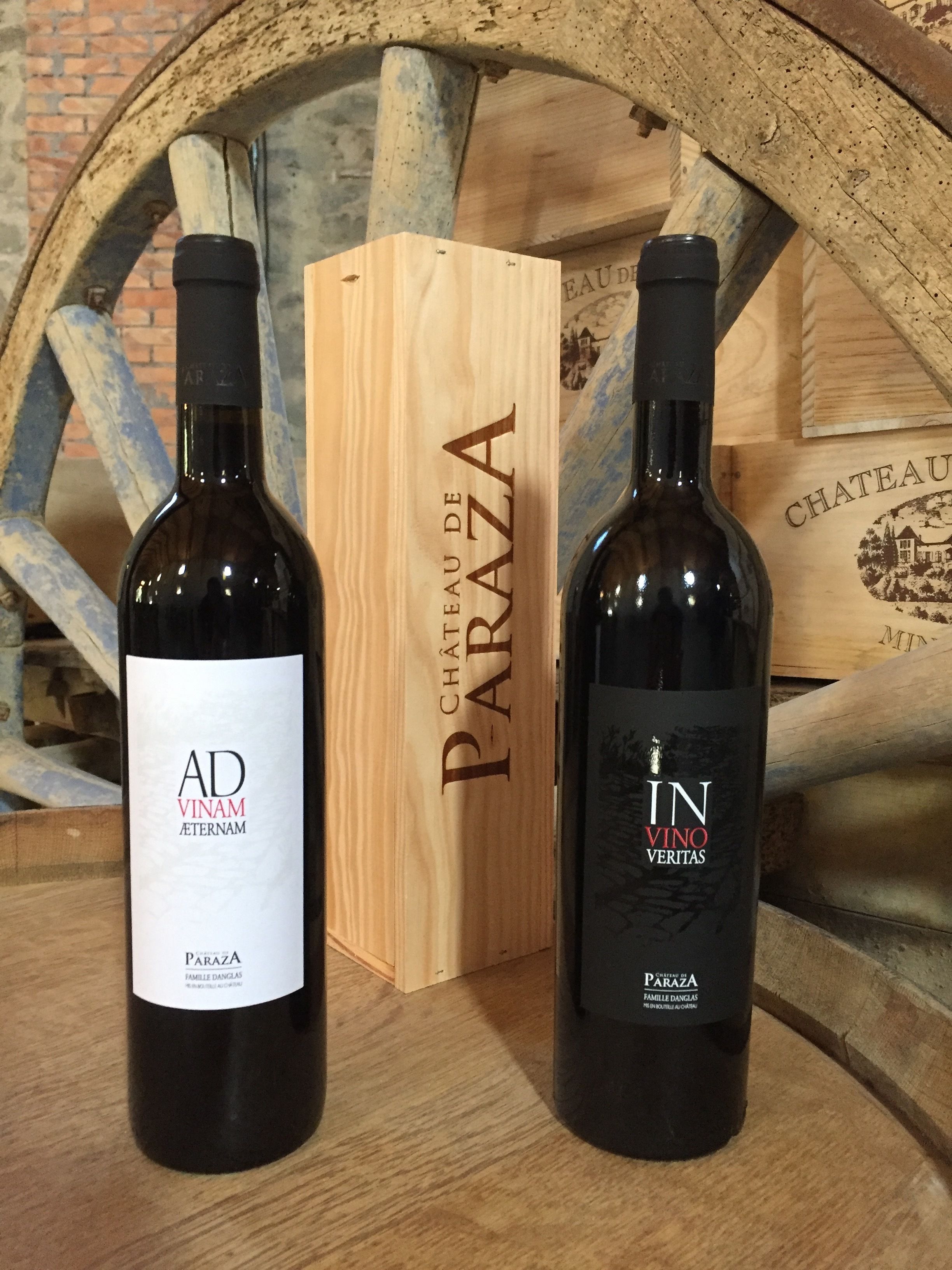 Dos botellas de vino etiquetadas 'AD VINAM' y 'IN VINO' sobre una superficie de madera, con una caja de madera y una rueda rústica al fondo.