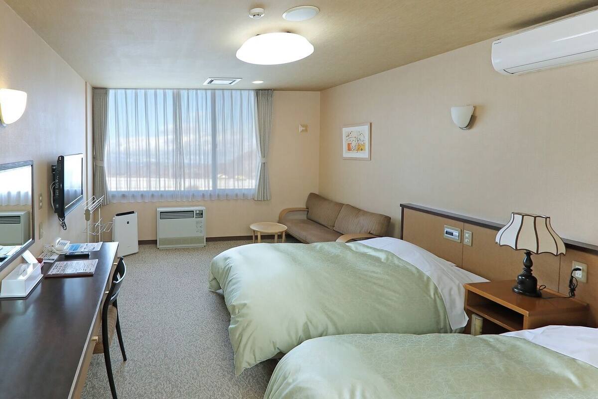 Habitación de hotel con escritorio, dos camas, un sofá, televisor, calefacción y una ventana con cortinas.