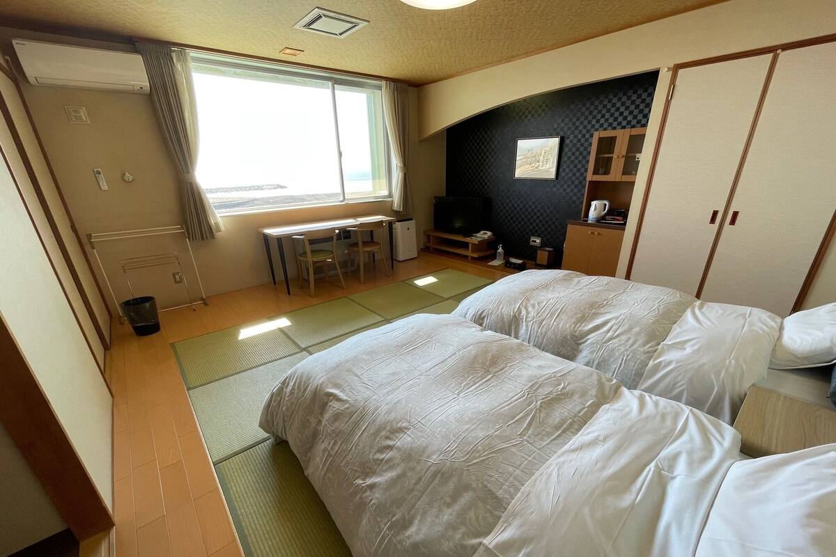 Habitación de hotel con esteras de tatami, una cama, un escritorio con sillas, un televisor y un armario.