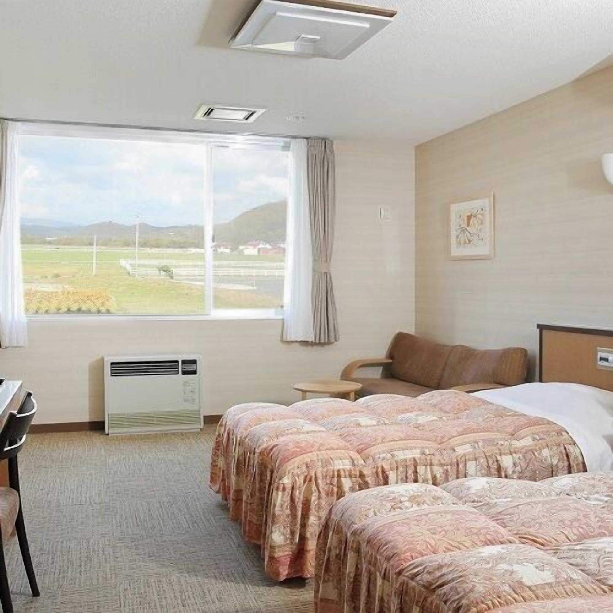 A hotel room with two beds, a desk, a chair, a window with a view of the countryside, and an air conditioning unit.