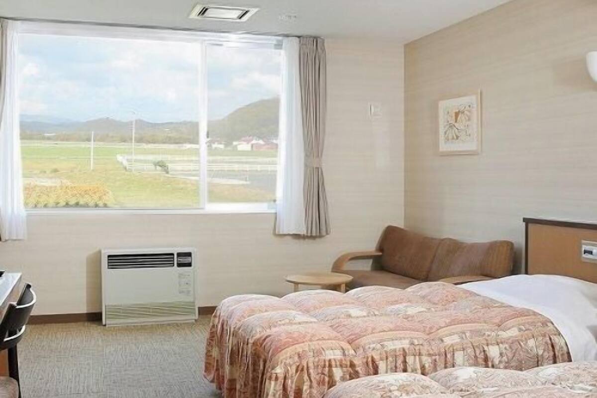 Habitación de hotel con dos camas, un escritorio, una silla, un ventanal con vistas al campo y un equipo de aire acondicionado.