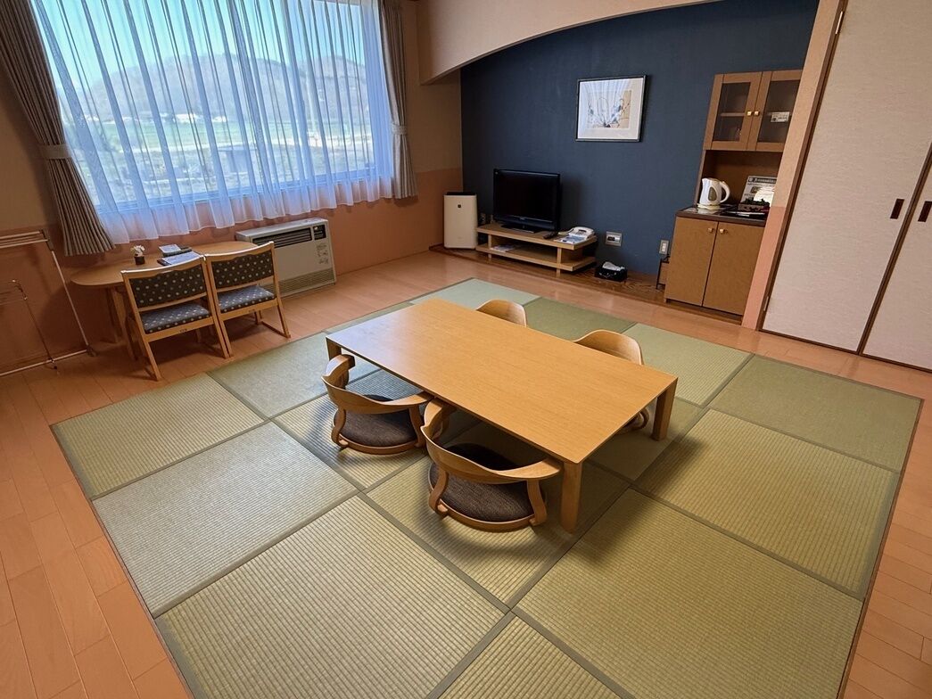 Una habitación con esteras de tatami, una mesa de madera, sillas, un televisor, un armario y una ventana con cortinas.