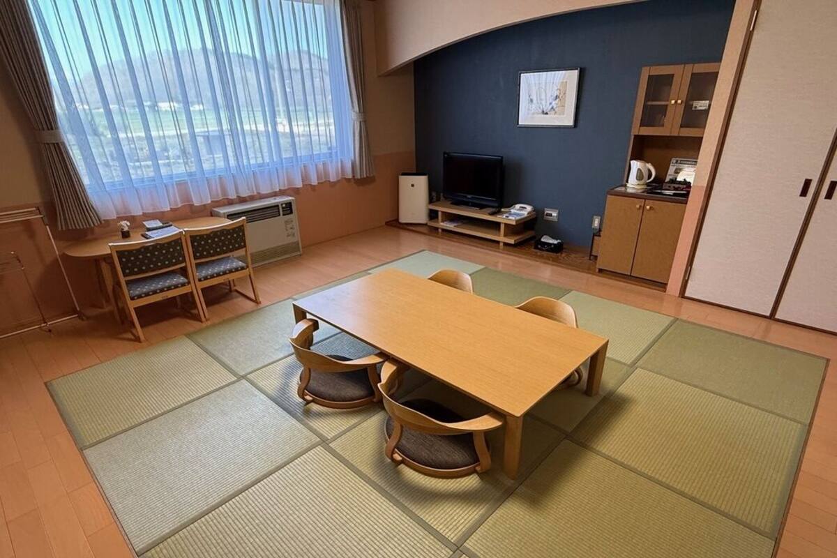 Una habitación con esteras de tatami, una mesa de madera, sillas, un televisor, un armario y una ventana con cortinas.
