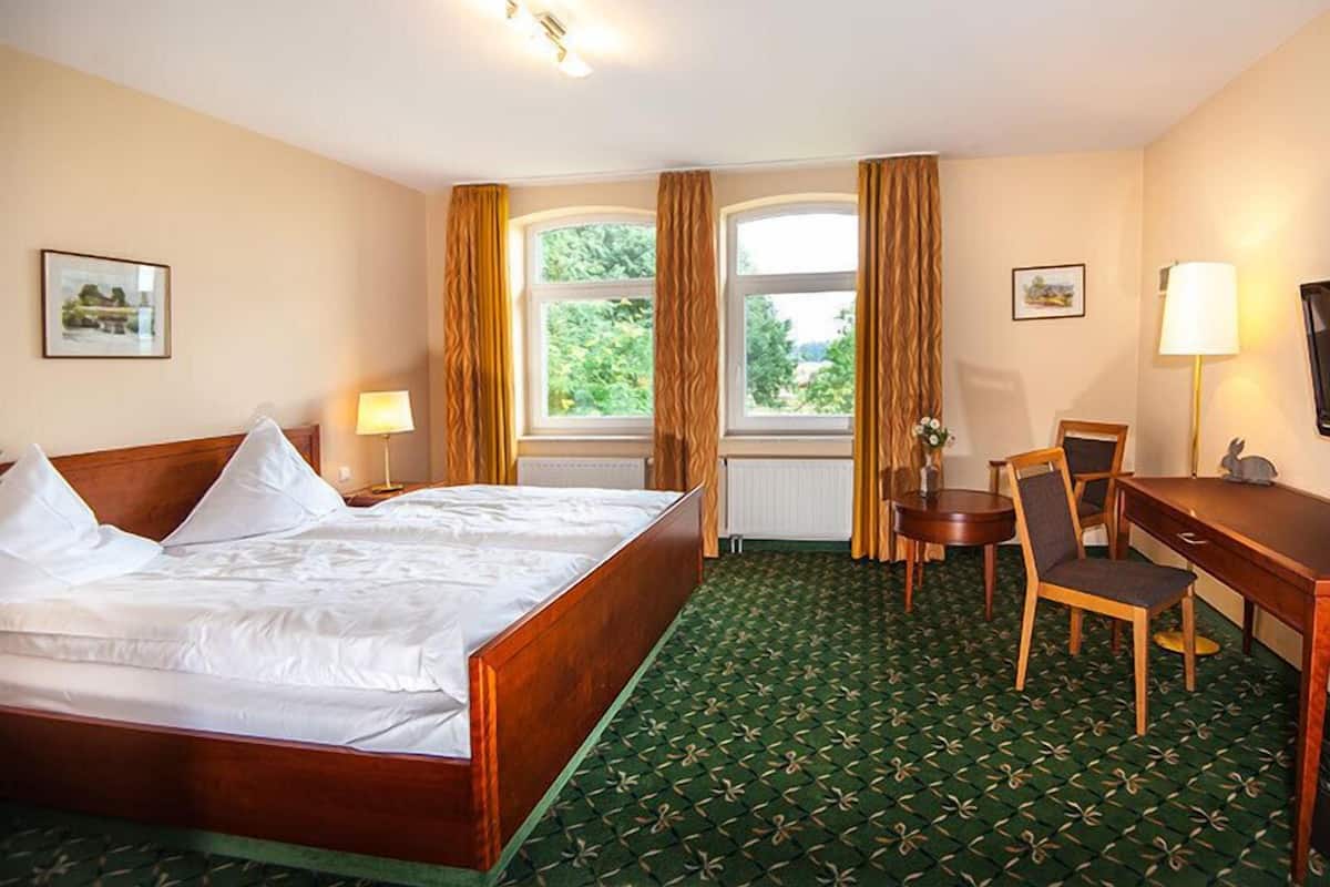 A hotel room with a wooden bed, a desk, a chair, and a view of greenery outside the window.