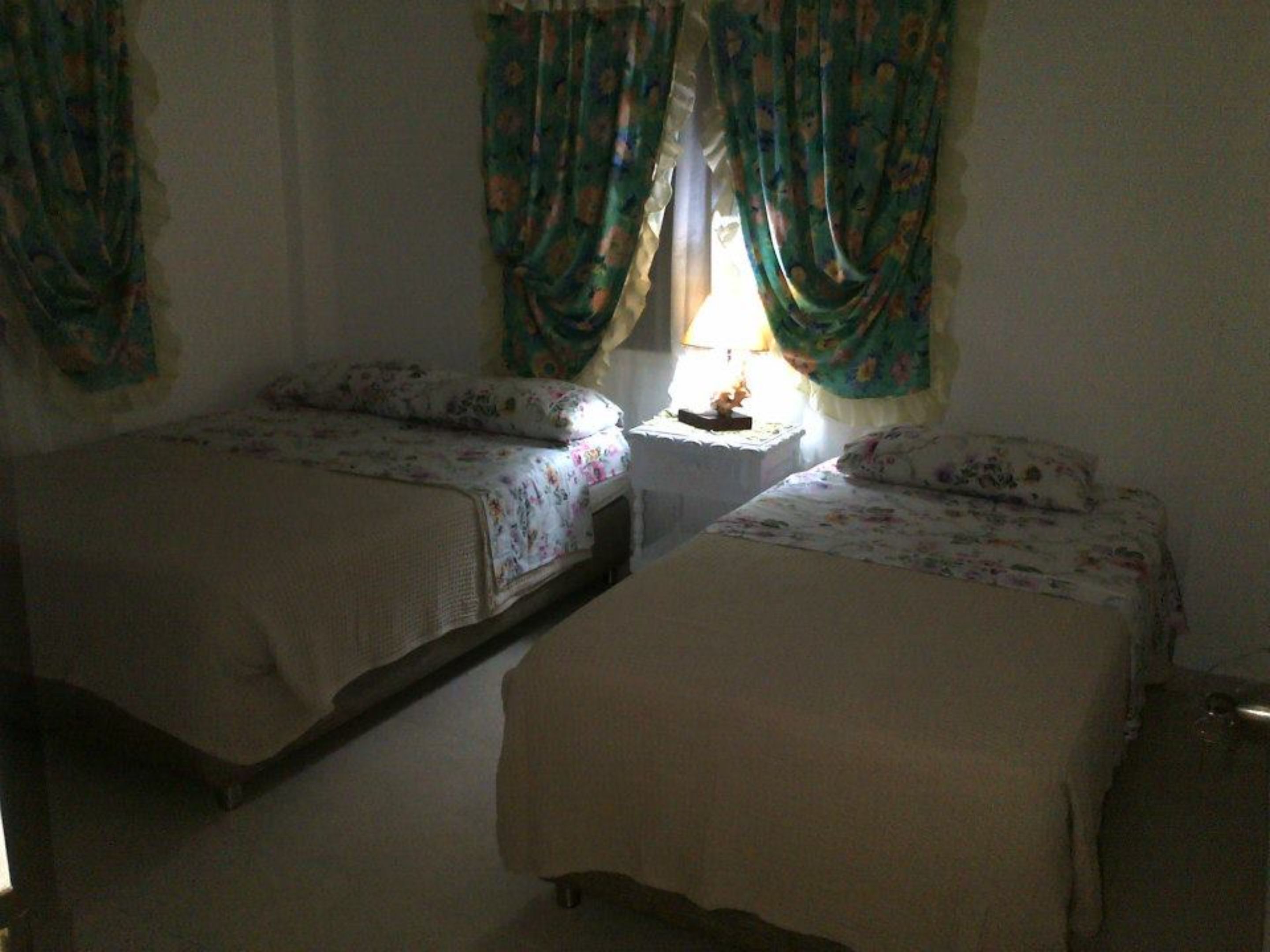 A small room with two single beds, a nightstand with a lamp, and floral curtains.