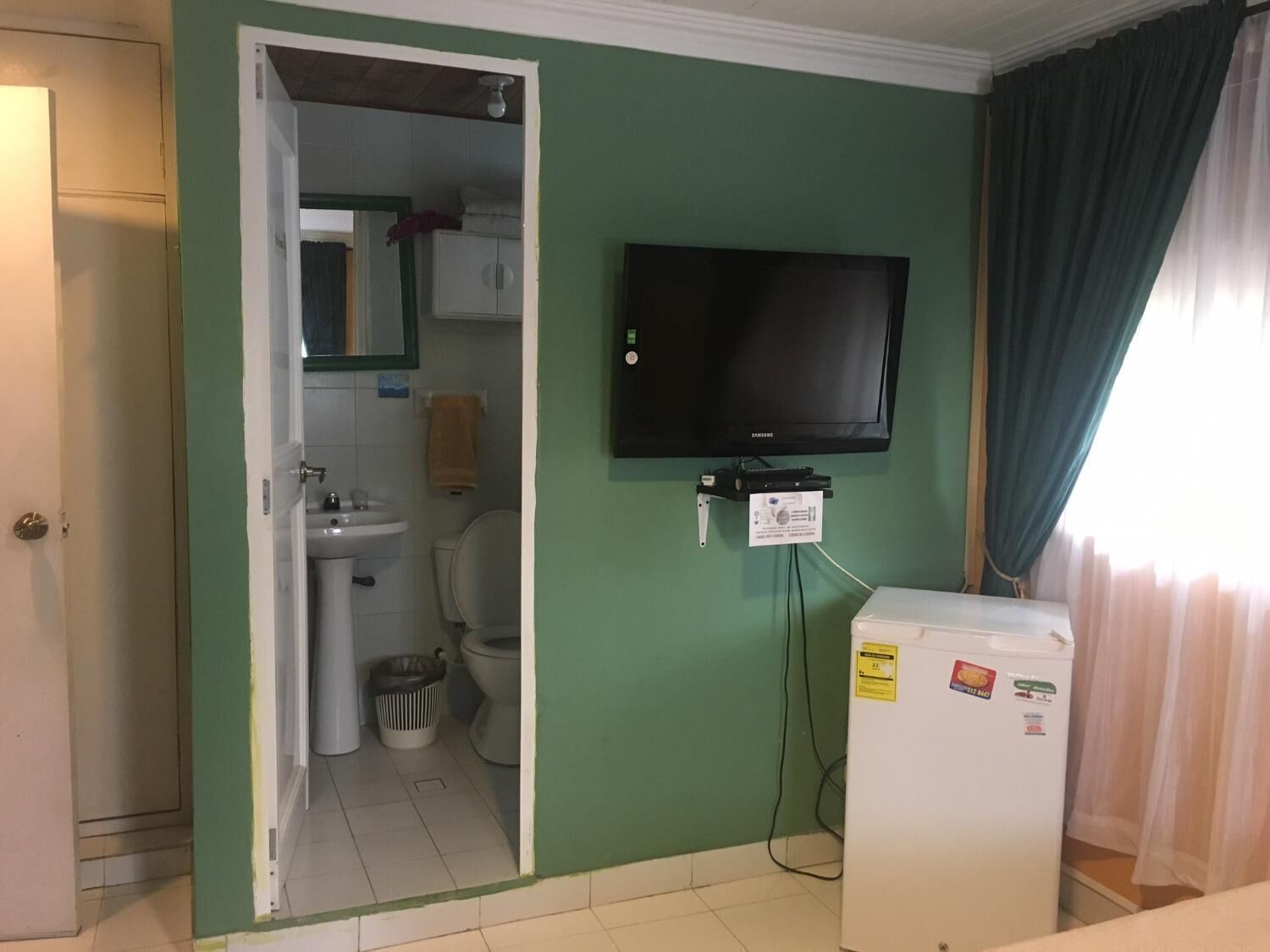 A room with a green wall, a mounted TV, a white refrigerator, a sink, and a toilet.