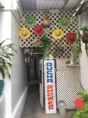 A narrow alleyway with a white fence, a sign reading "DUCHA SHOWER", and various colorful woven hats hanging above.