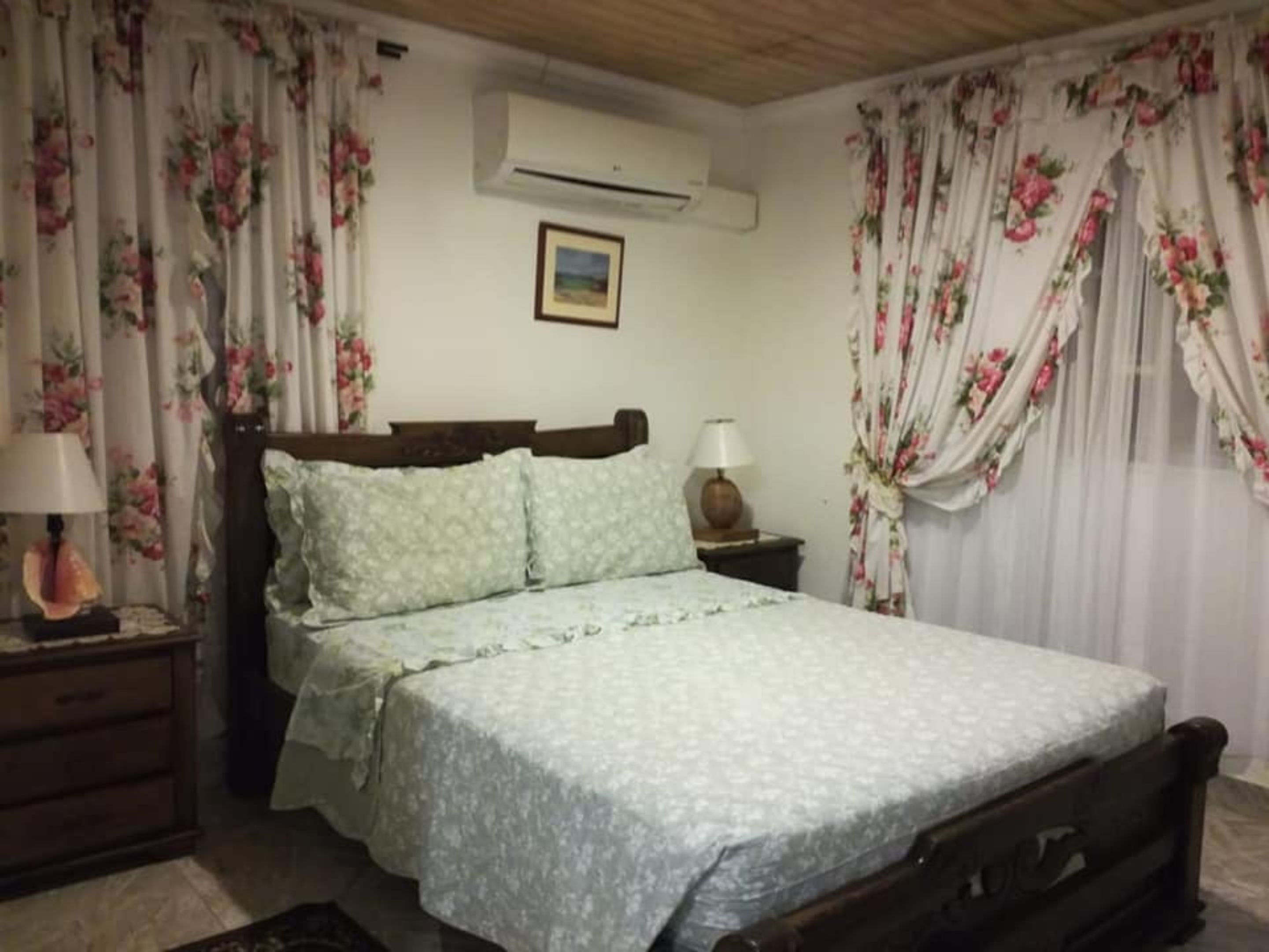 A bedroom with a bed, a nightstand, a lamp, floral curtains, and an air conditioning unit.