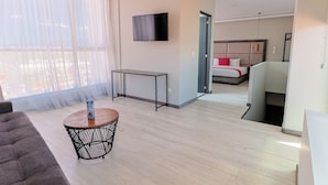 Quarto de hotel moderno com uma grande janela, uma televisão de tela plana, uma pequena mesa redonda e uma cama com almofada vermelha decorativa.