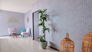 Um quarto de spa com uma grande planta em vaso, duas cadeiras de vime, uma mesa pequena e uma placa indicando 'Airkana Spa'.