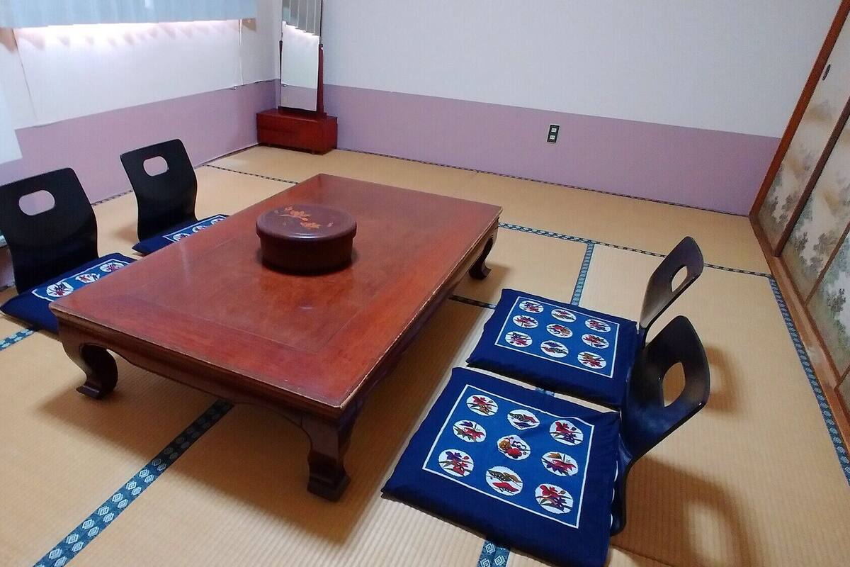 Una stanza con pavimento in tatami, un tavolo in legno e sedie, un piccolo armadio e uno specchio a parete.