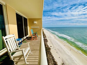 A balcony with a view of a beach, featuring a white rocking chair, a blue chair, and a small table.