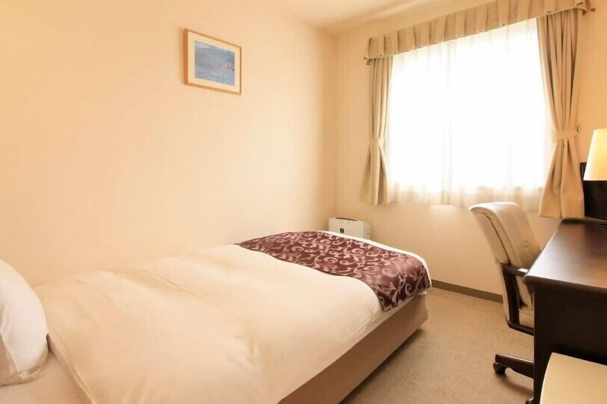 A hotel room with a bed, a desk, a chair, a window with curtains, and a picture on the wall.
