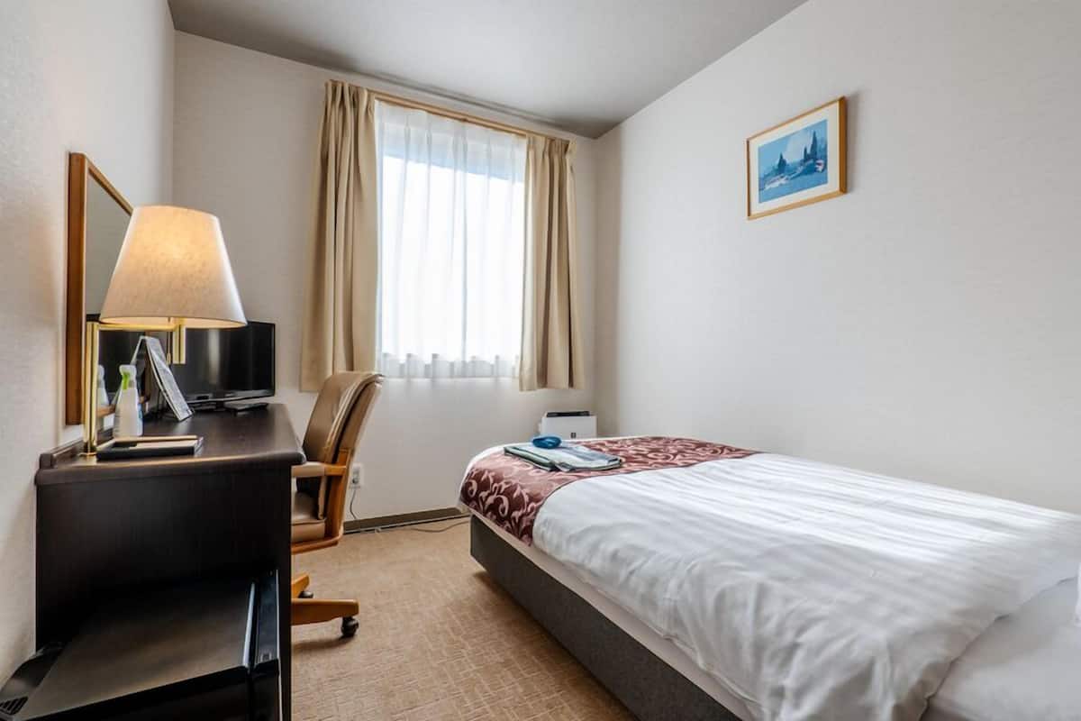 A hotel room with a bed, a desk with a lamp, a chair, a window with curtains, and a framed picture on the wall.