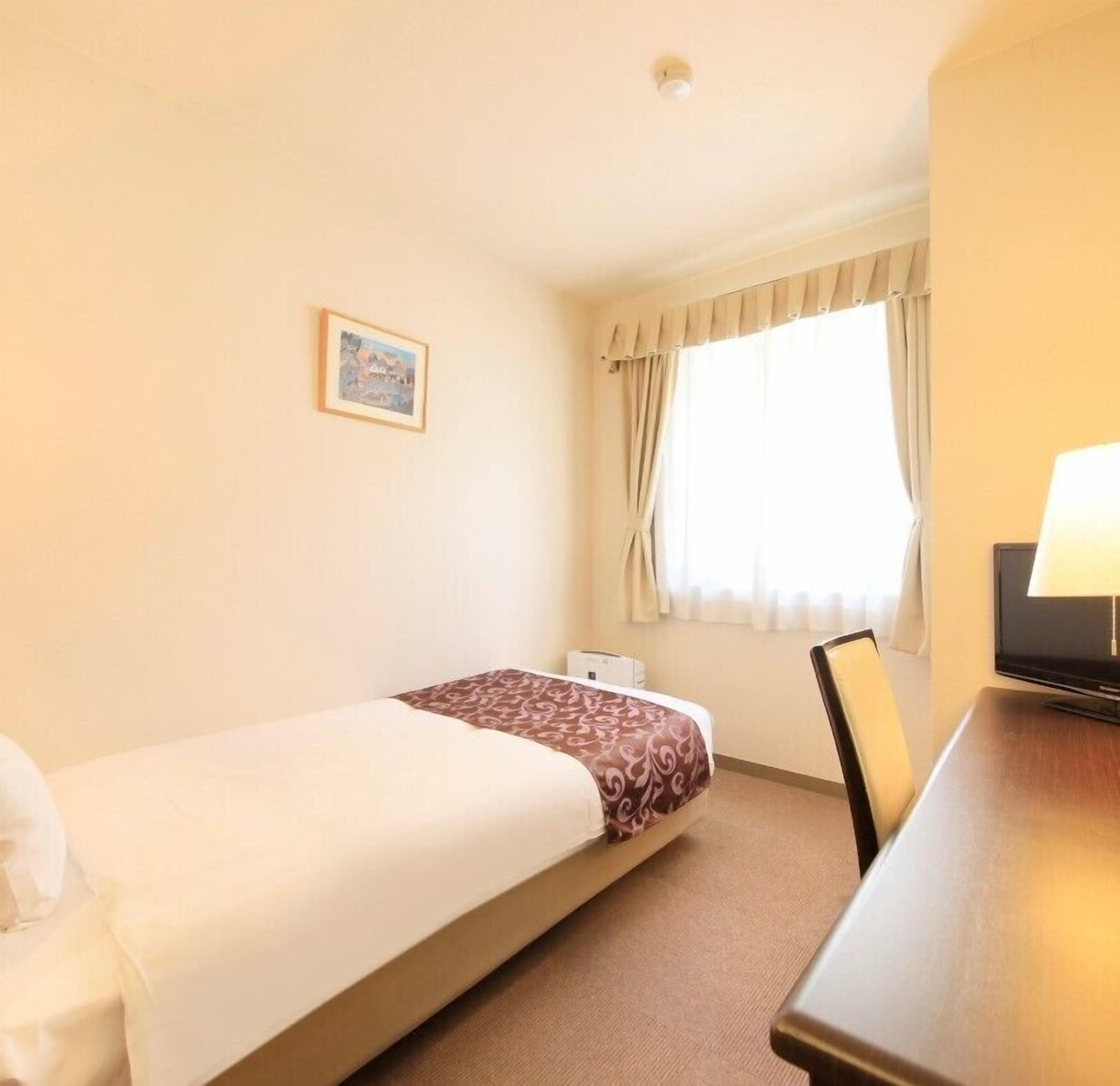 Ein Hotelzimmer mit einem Einzelbett, einem Schreibtisch, einem Stuhl, einem Fernseher und einem Fenster mit Vorhängen.