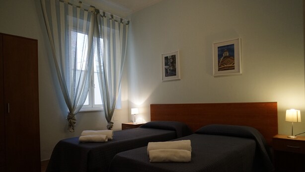A hotel room with two single beds, a wooden wardrobe, a window with sheer curtains, and two framed pictures on the wall. - Casa Cappone (Alghero)
