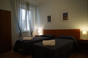 Habitación de hotel con dos camas individuales, un armario de madera, una ventana con cortinas traslúcidas y dos cuadros en la pared.