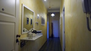 A narrow hallway with yellow walls, a white door, and a white-framed mirror. There are framed pictures on the wall, a small shelf with a vase of flowers, and a hanging light fixture. - Casa Cappone (Alghero)