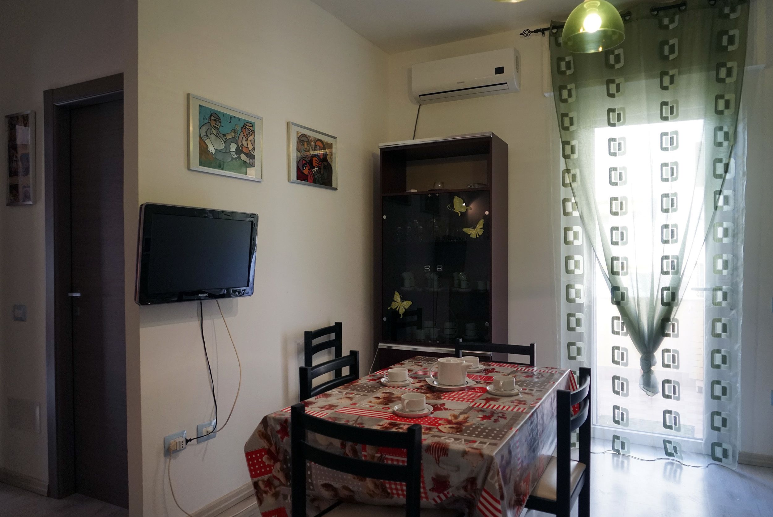 Un comedor con una mesa dispuesta para cuatro, un televisor de pantalla plana montado en la pared y un aparador con puertas de cristal.