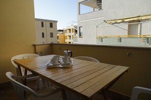 Un balcón con mesa de madera y sillas de plástico blanco, una cafetera y una bandeja con tazas y platillos.